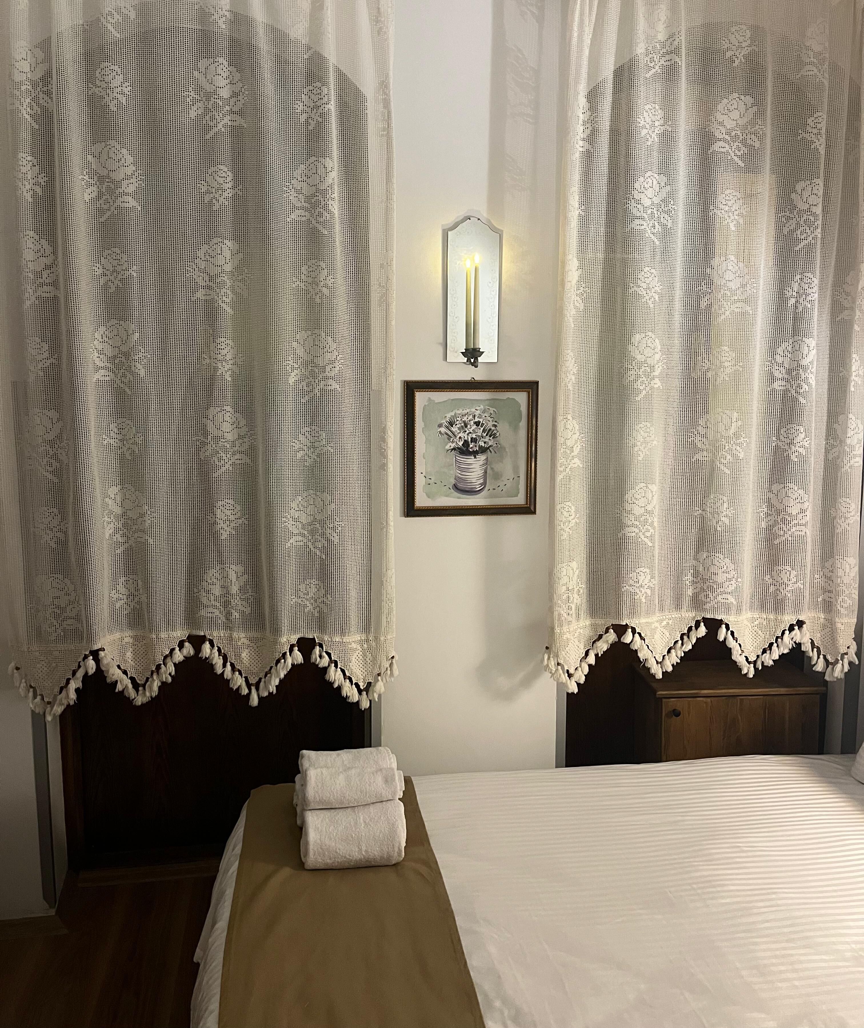 A hotel room with a bed, two nightstands, a framed picture, and lace curtains.