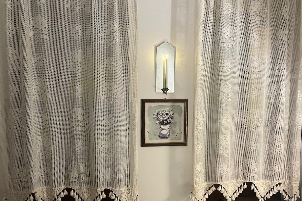 Habitación de hotel con una cama, dos mesitas de noche, un cuadro enmarco y cortinas de encaje.
