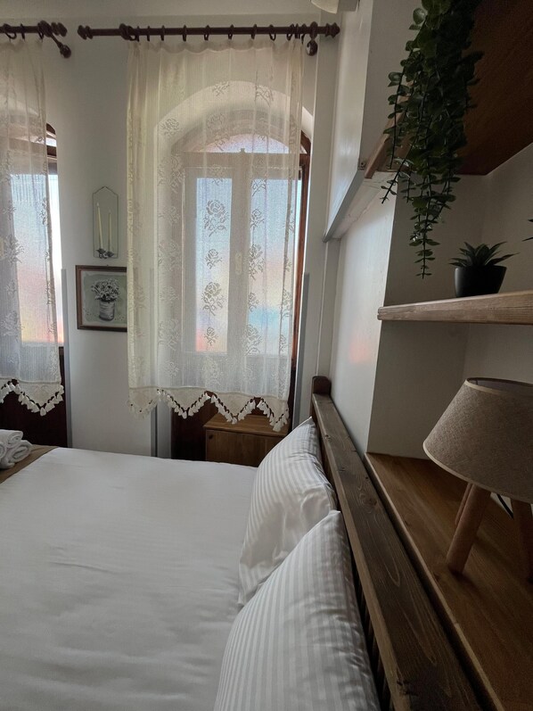 A bedroom with a bed, a nightstand, a lamp, a potted plant, and a window with lace curtains.
