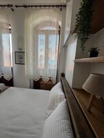 Un dormitorio con cama, mesita de noche, lámpara, planta en maceta y ventana con cortinas de encaje.