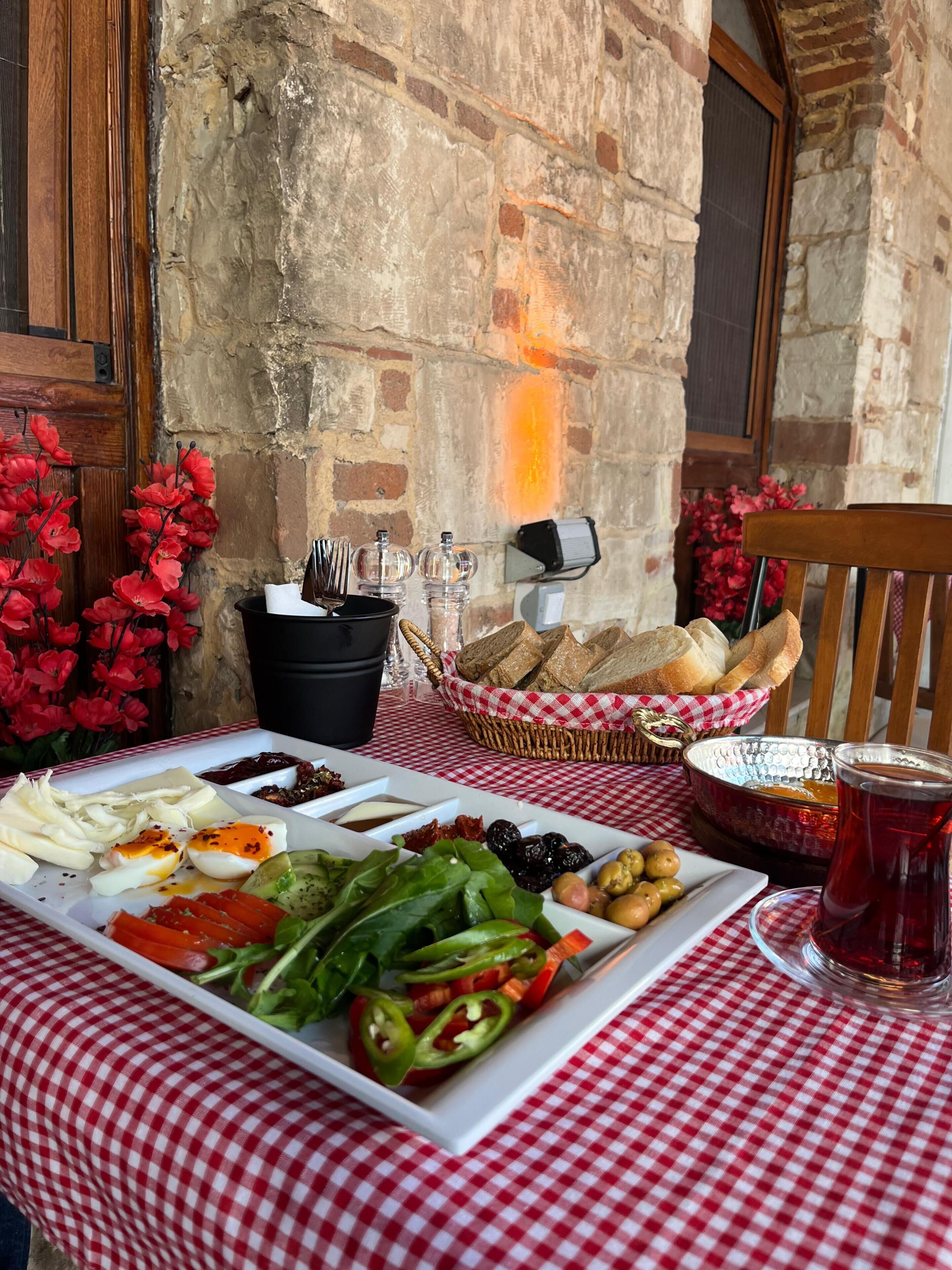 Ein Tisch ist reichhaltig mit Speisen gedeckt, darunter ein Brotkorb und ein Glas rotes Getränk, vermutlich Tee, im Freien vor einer Ziegelwand mit roten Blumen im Hintergrund.