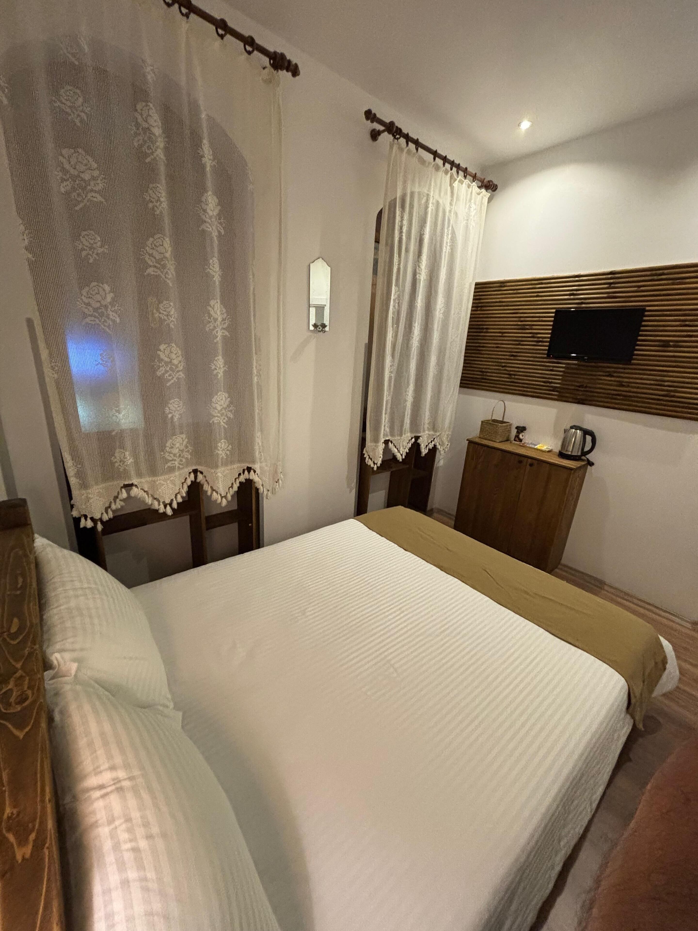 Ein Hotelzimmer mit Bett, einem Nachttisch aus Holz, einem an einer Holzplatte montierten Fernseher und einem Fenster mit durchsichtigen Vorhängen.
