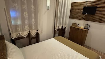 Habitación de hotel con cama, mesita de noche de madera, televisor montado en un panel de madera y una ventana con cortinas de gasa.