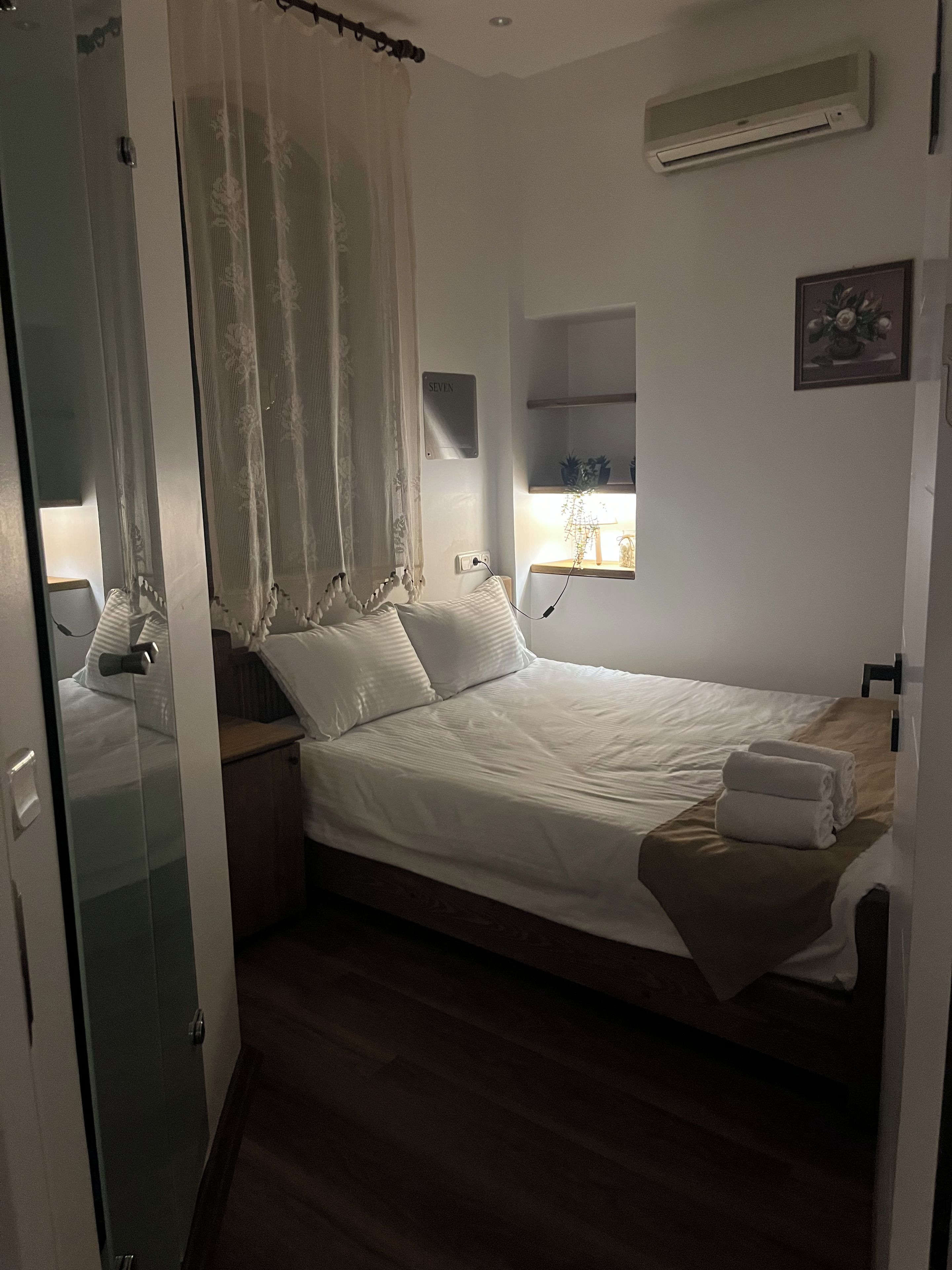 Un dormitorio con cama, mesita de noche, ventana con planta, aire acondicionado de pared y puerta con panel de vidrio.