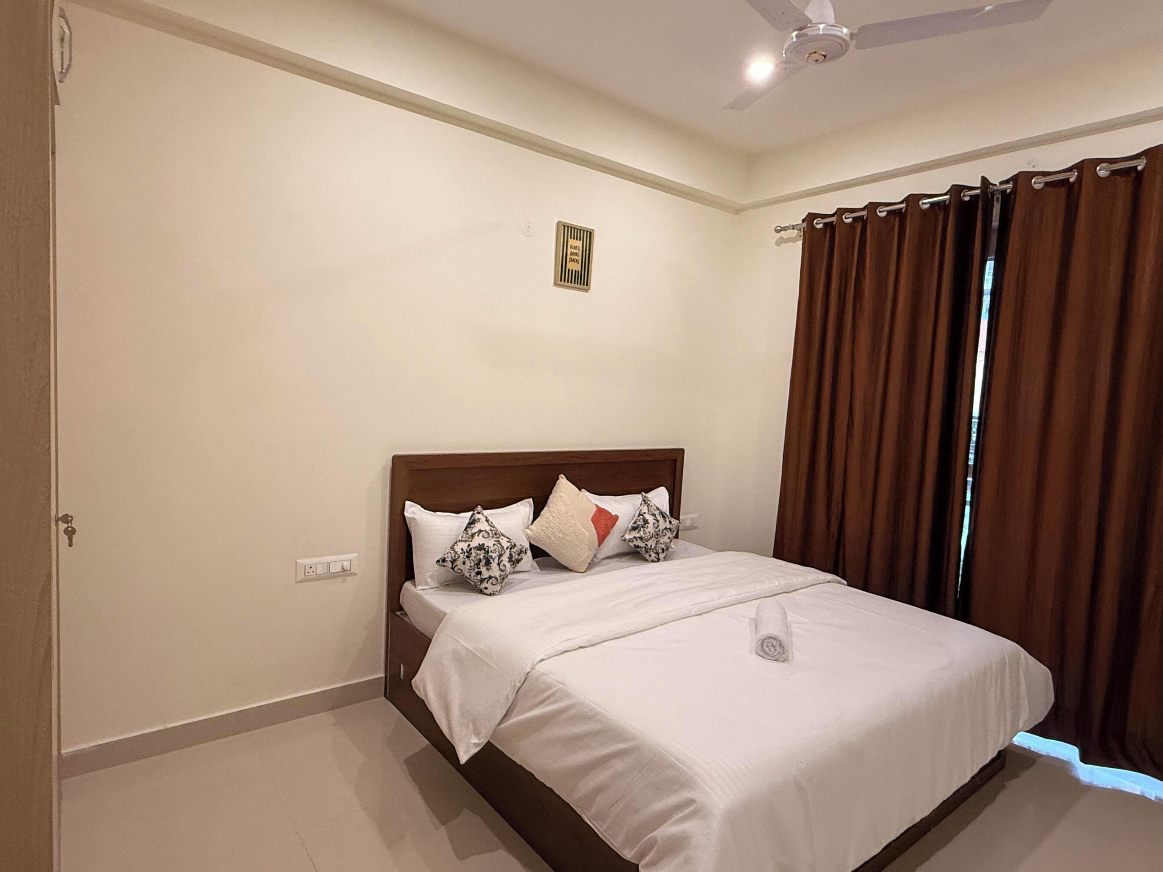 A hotel room with a bed, a ceiling fan, a window with curtains, and a wall-mounted light switch.