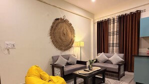 Una sala de estar con dos sofás, un sillón de peluche amarillo, una mesita de centro, un tapiz en la pared y una cortina.