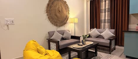 A living room with two sofas, a yellow bean bag, a coffee table, a wall hanging, and a curtain.