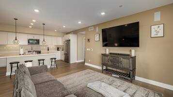 A modern living room with a large flat-screen TV, a sofa, and a kitchen in the background.