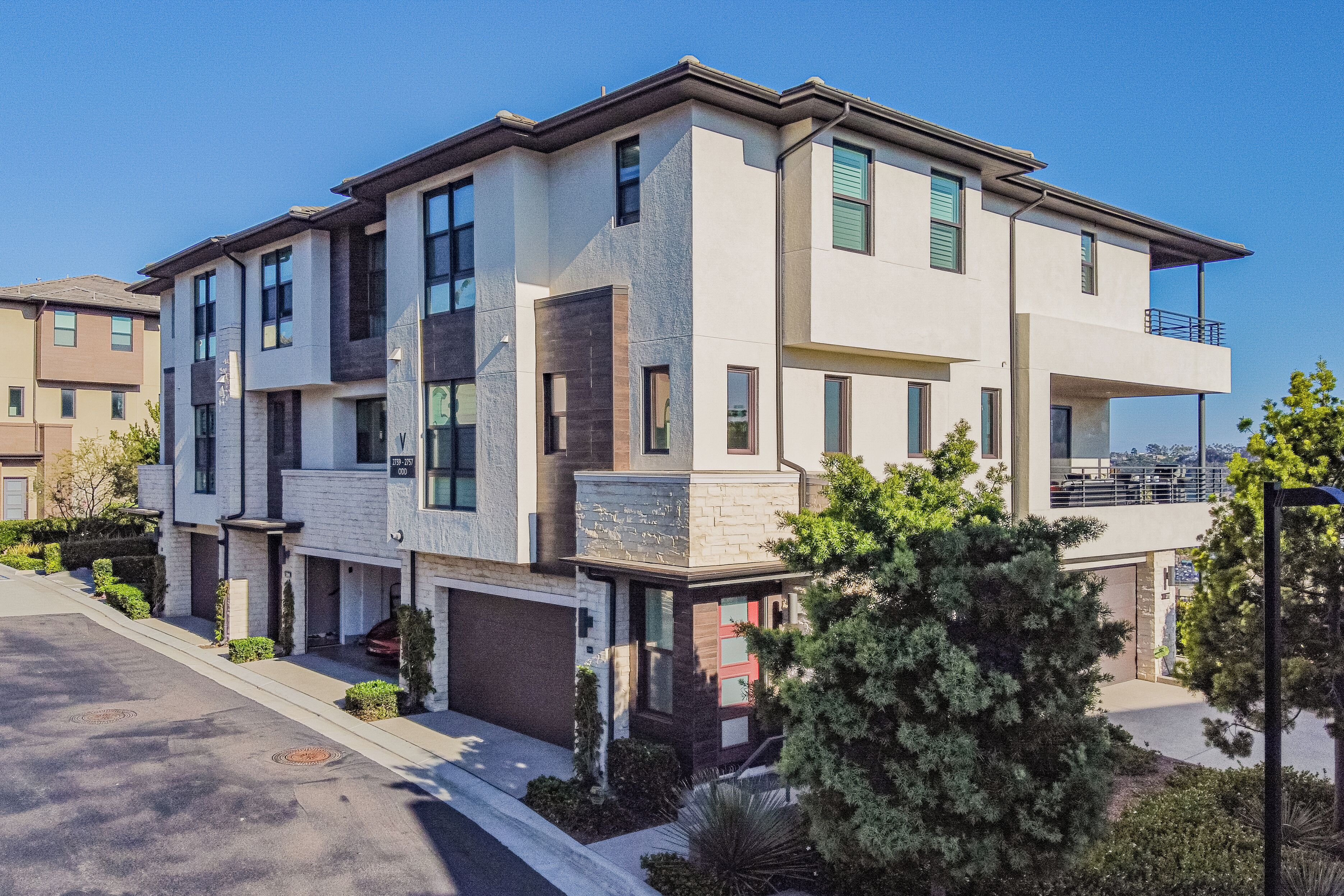 A modern multi-story residential building with a garage, a balcony on the upper floor, and a well-maintained front yard with landscaping.
