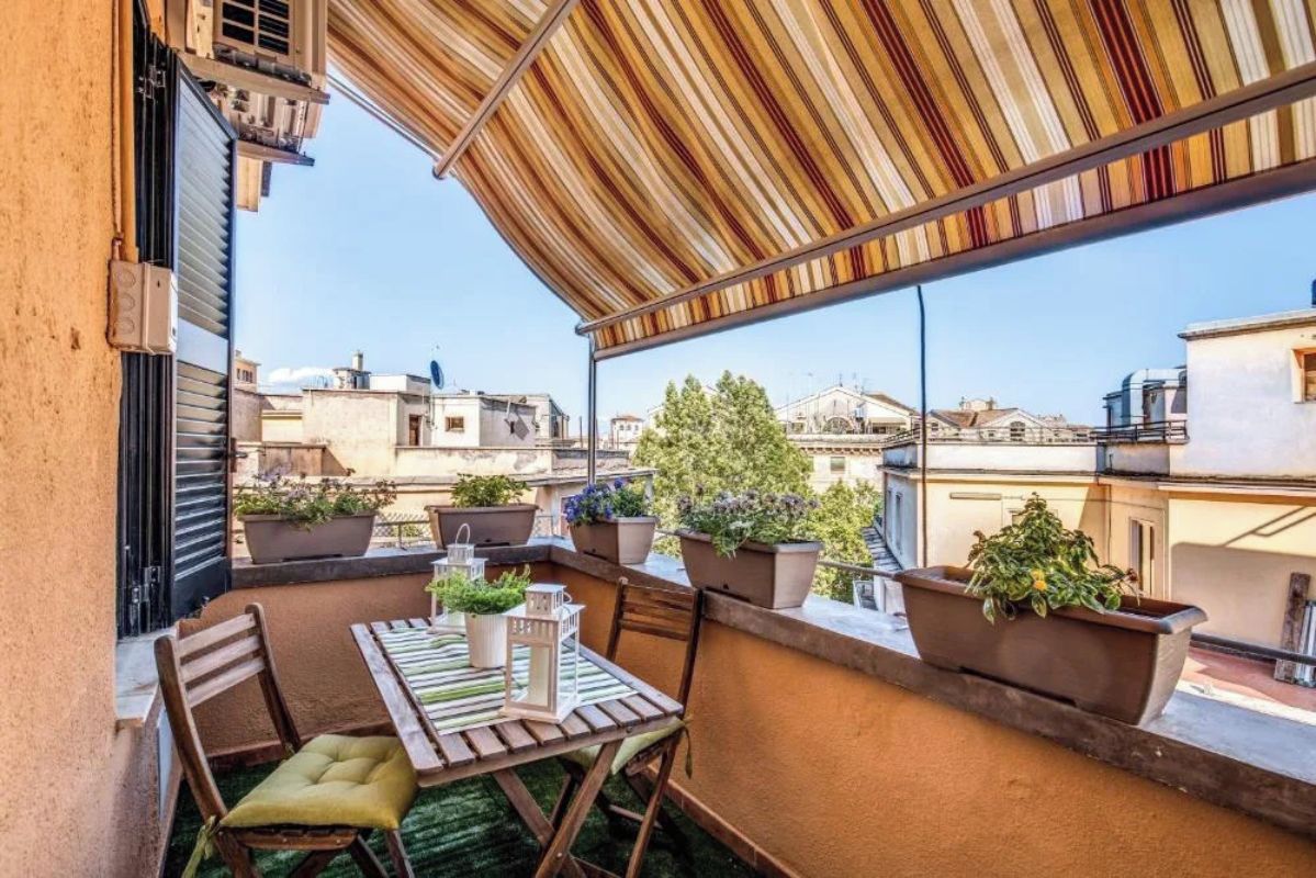 Un balcon agrémenté d’un auvent à rayures, de plantes en pot, d’une table avec des chaises et offrant une vue sur le paysage urbain.
