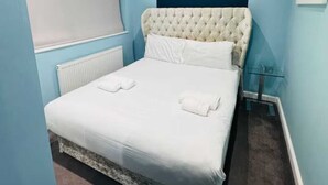Un dormitorio con cabecera capitonada, una cama grande con ropa blanca, un radiador y una ventana con persiana.