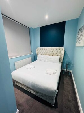 A bedroom with a tufted headboard, a large bed with white linens, a radiator, and a window with a blind. - Milburn Residents (Oldbrook)