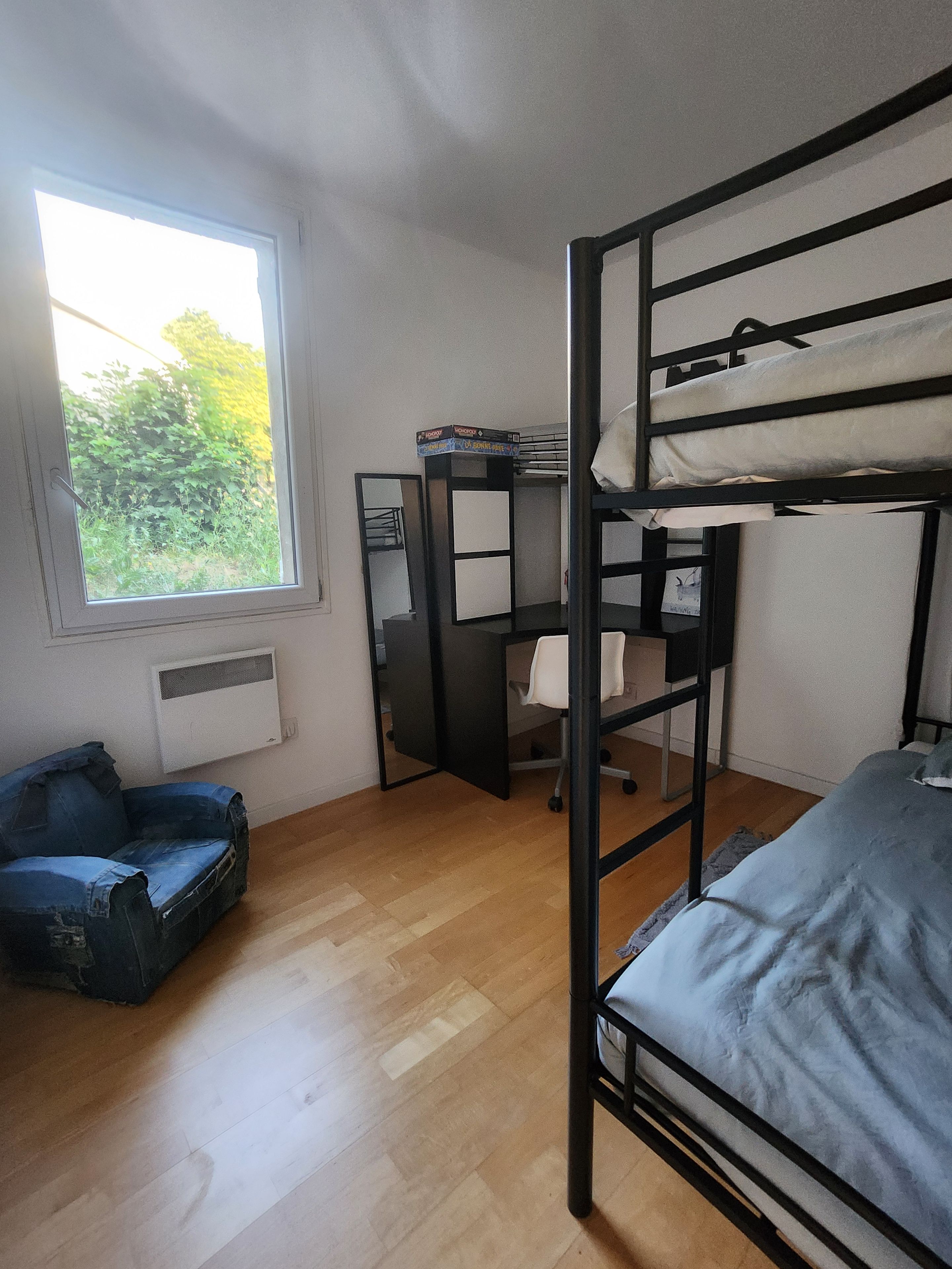 Une chambre avec un lit superposé, un bureau avec une chaise, un radiateur et une fenêtre donnant sur un espace verdoyant.