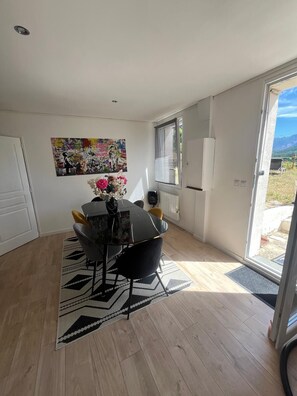 A dining area with a table and chairs, a patterned rug, a wall-mounted artwork, and a window with blinds. - Beautiful 130-square-meter house with a view of the mountains (Méreuil)