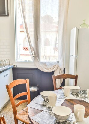 A dining area with a table set for four, chairs, a window with curtains, and a refrigerator. - Spacious 3-bedroom apartment in delightful Cannes with AC - LES BANANIERS (Cannes)