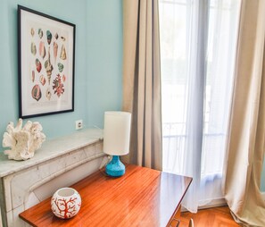 Una habitación con una mesa de madera, un jarrón blanco con diseño de árbol rojo, una lámpara azul y un cuadro con caracoles en la pared.
