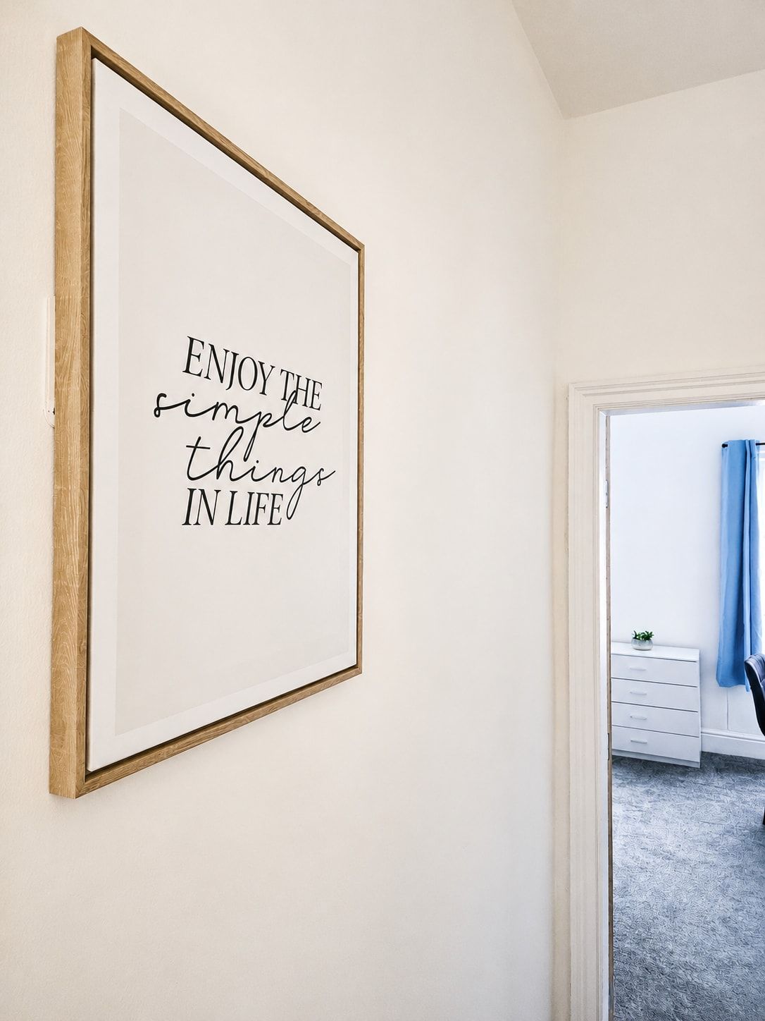 A framed inspirational quote on a wall in a room with a white dresser and a blue curtain.