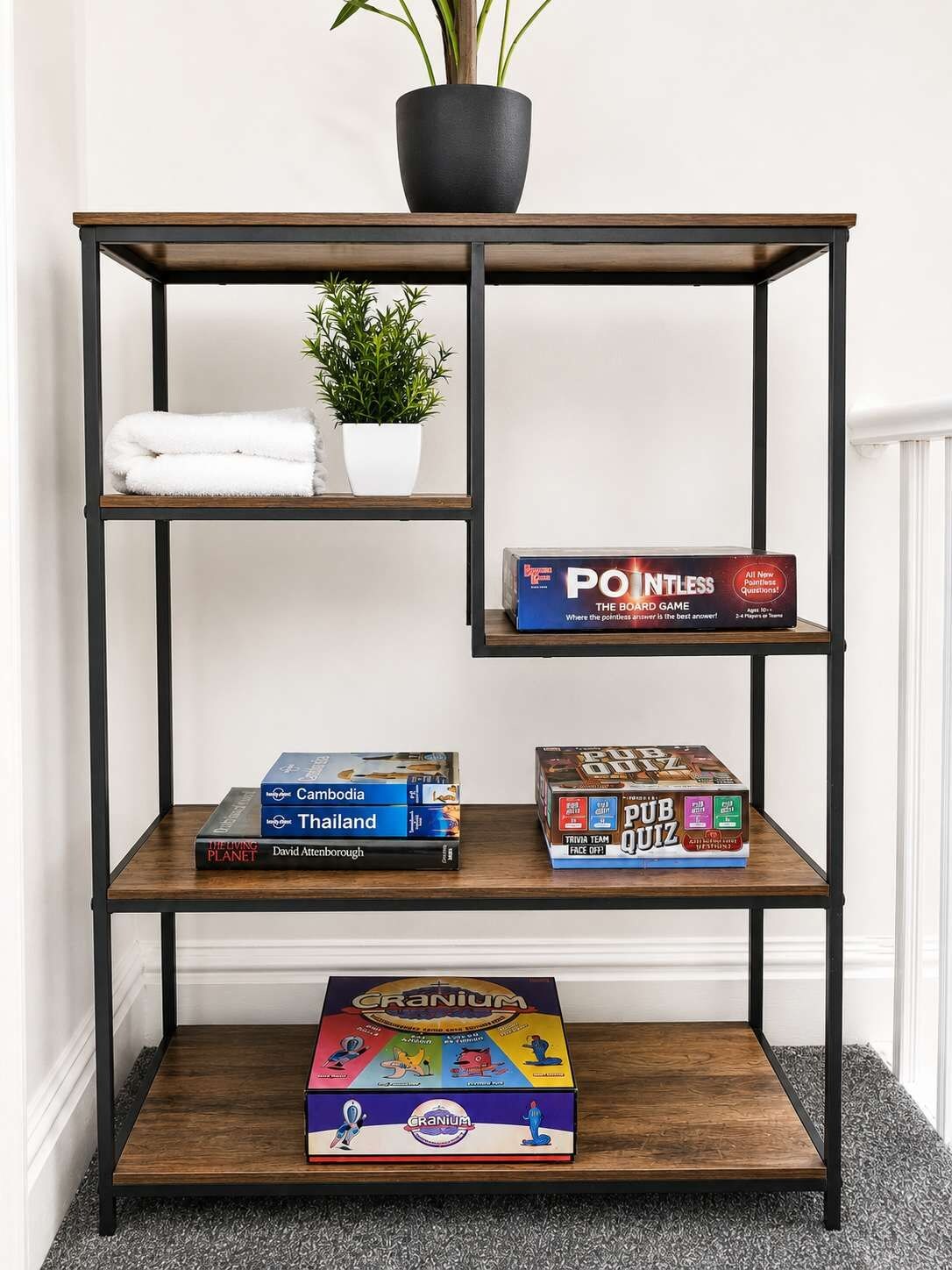 A wooden bookshelf with three shelves, holding books, a board game, and a potted plant.