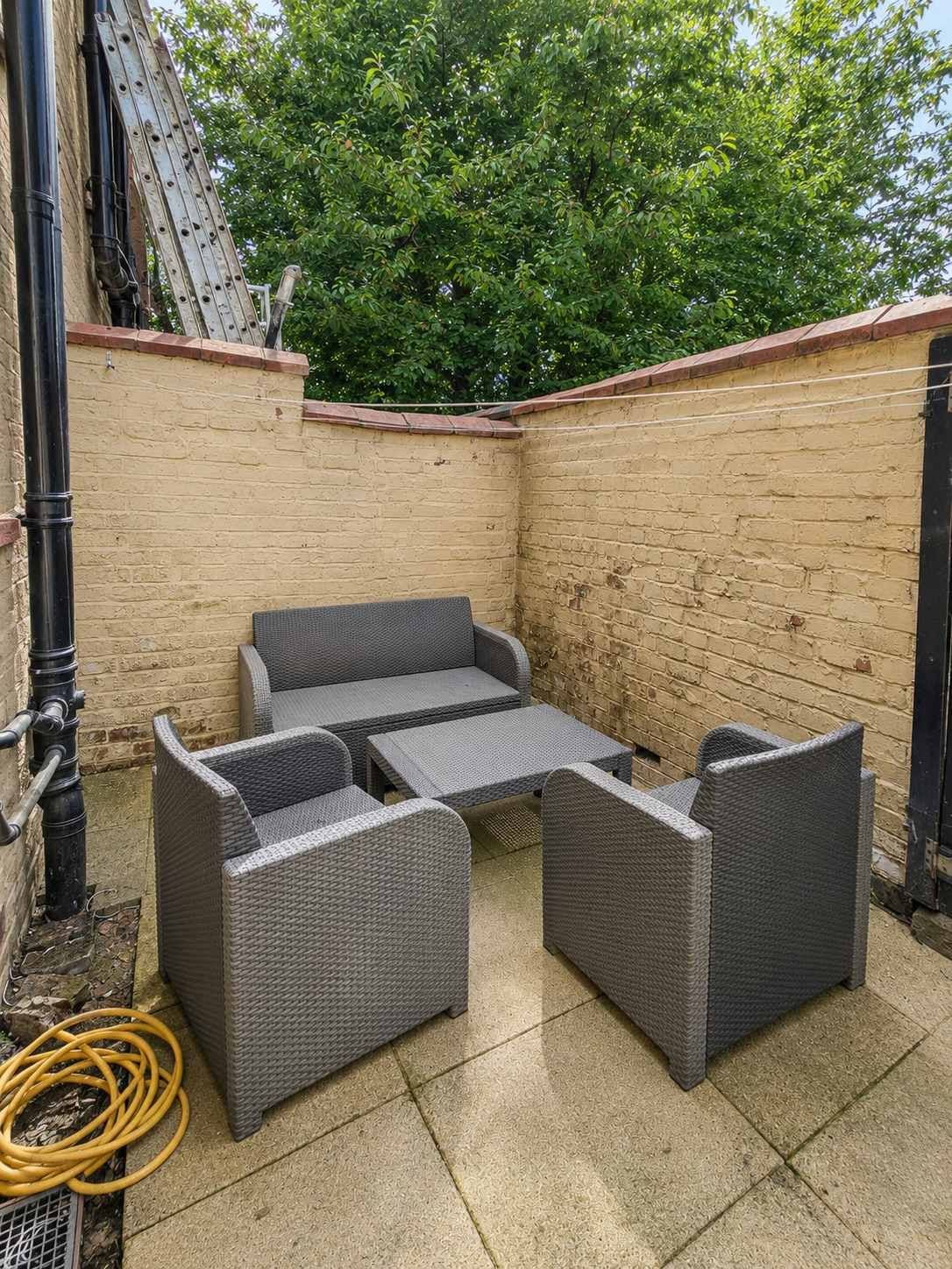 Un espace patio avec une table et des chaises en osier, un mur de briques et un arbre en arrière-plan.