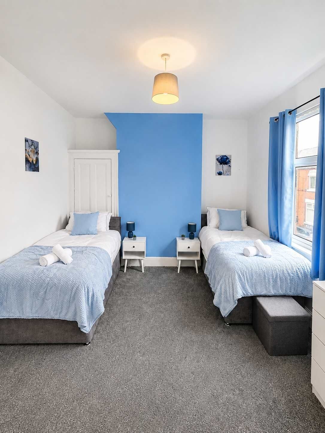A bedroom with two beds, a white door, a blue accent wall, a ceiling light, and a window with curtains.