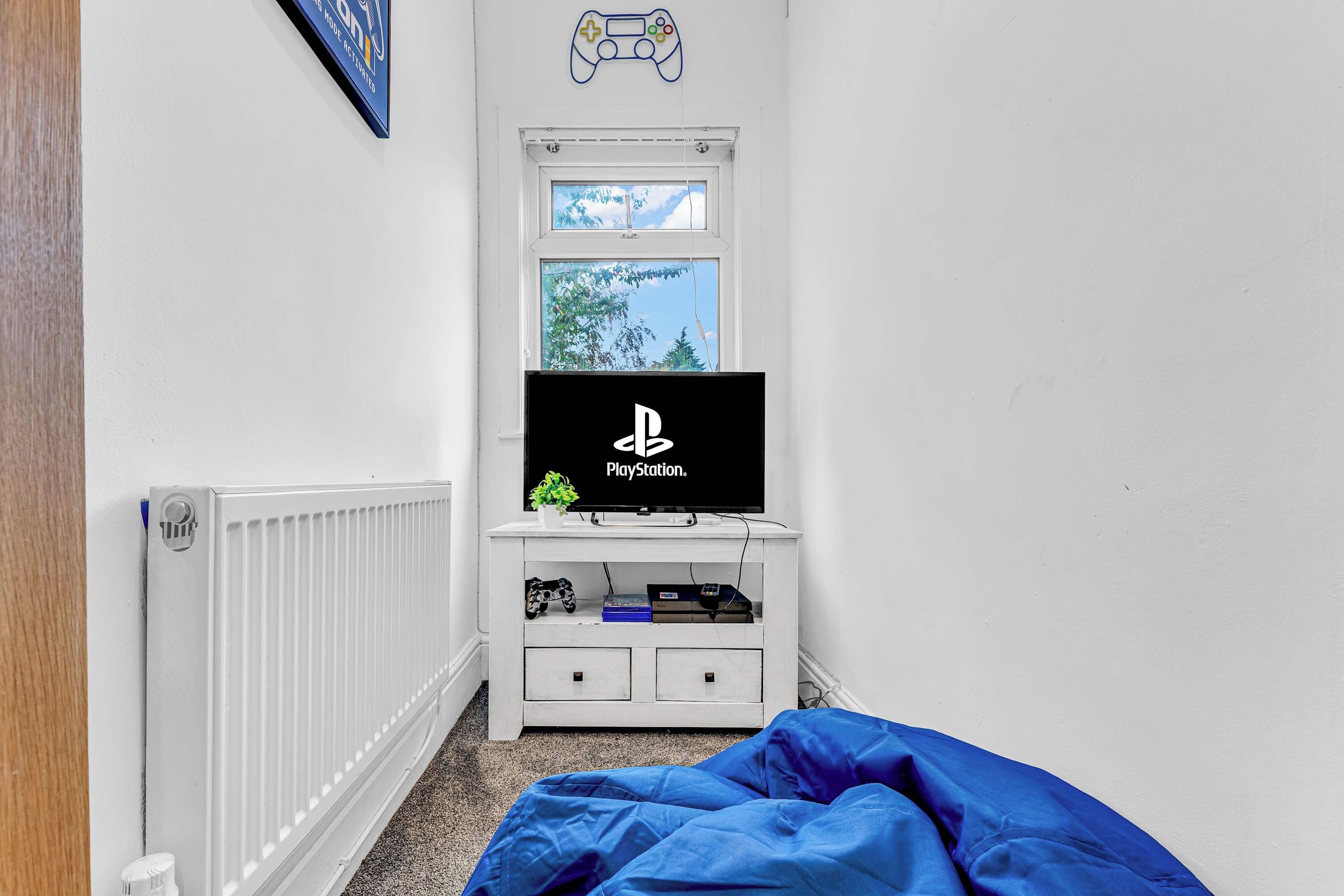 A room with a white radiator, a PlayStation TV on a white stand, a window with a view of trees, and a blue blanket on the floor.