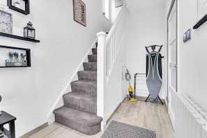 A hallway with a carpeted staircase, a black and white patterned rug, and a wall-mounted ironing board. - Modern 3 Bed Home | Driveway | Spacious Garden (Stretford)