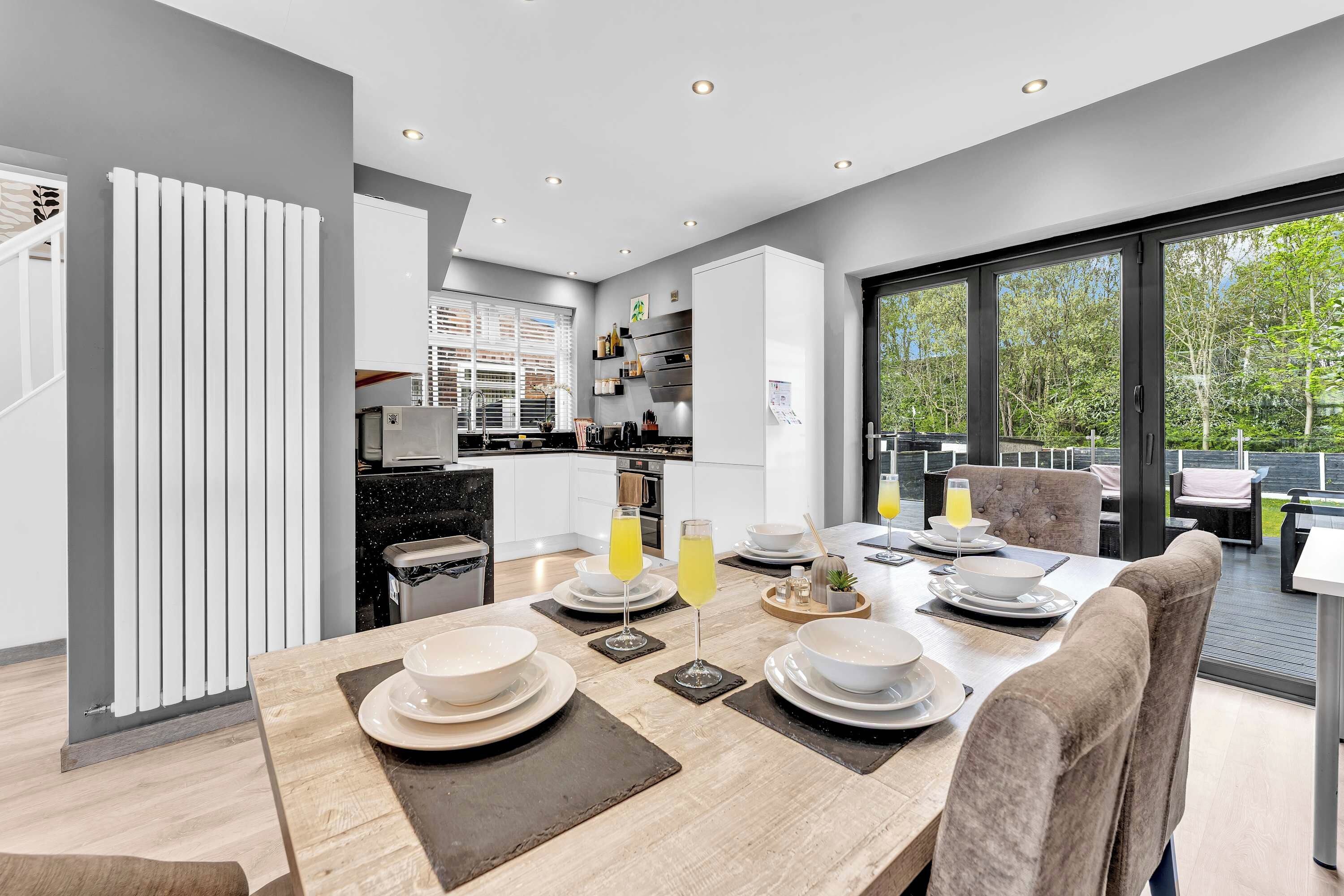 A modern kitchen with a dining area, large windows, and a view of greenery.
