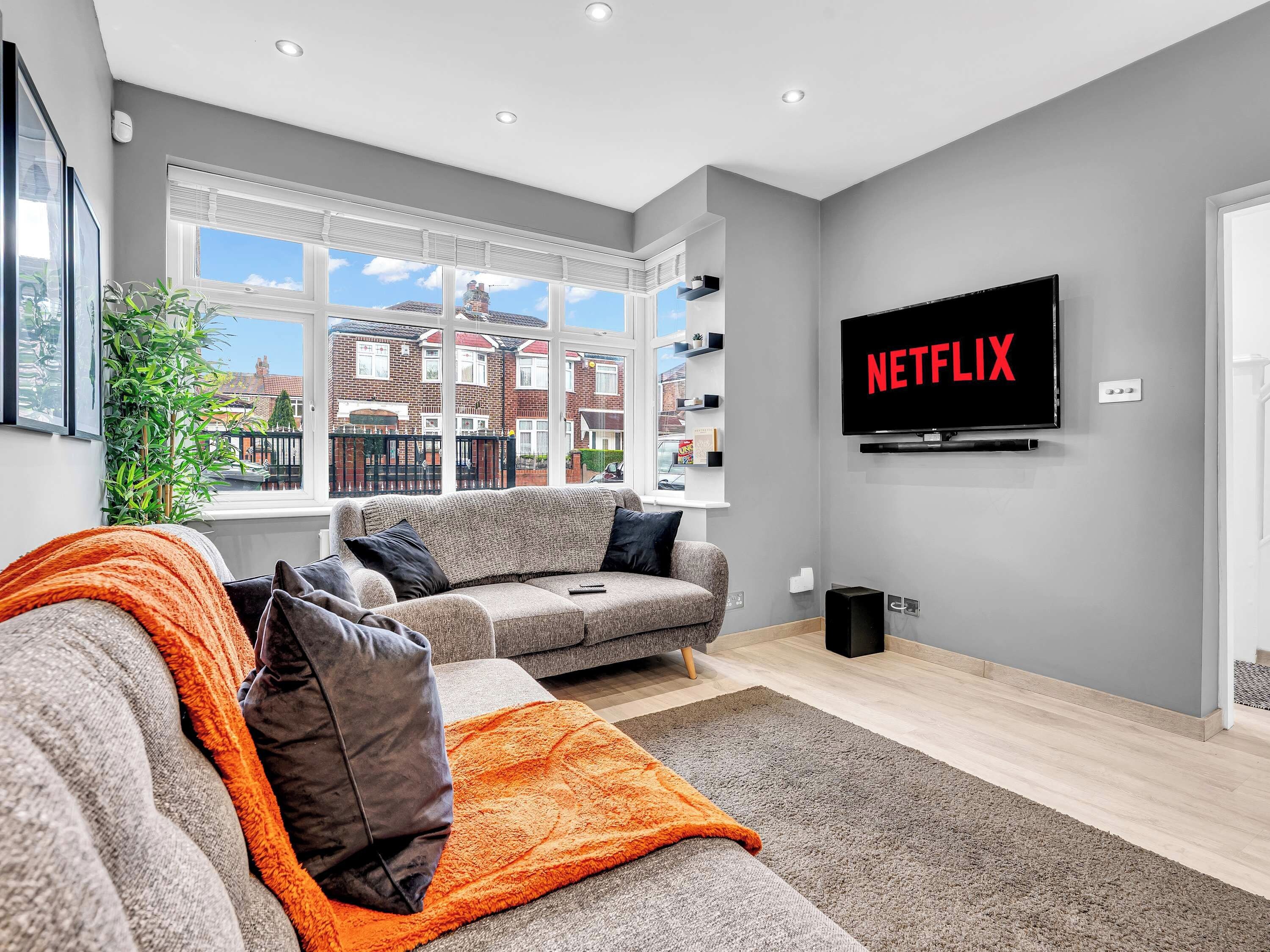 Een moderne woonkamer met een grijze bank, een televisie waarop 'NETFLIX' wordt weergegeven en een groot raam met uitzicht op een woonwijk.