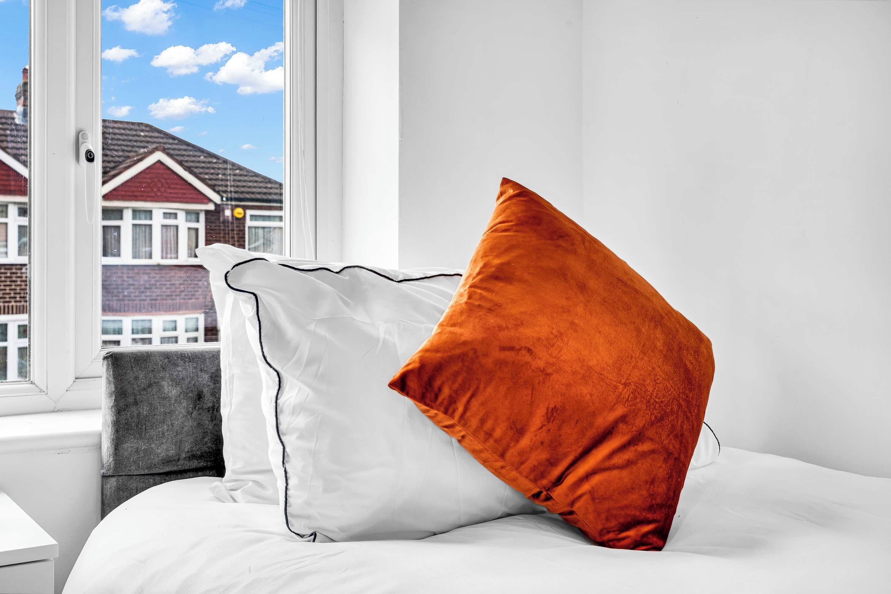 A bed with white bedding and two pillows, one white and one orange, in a room with a window overlooking a residential area.