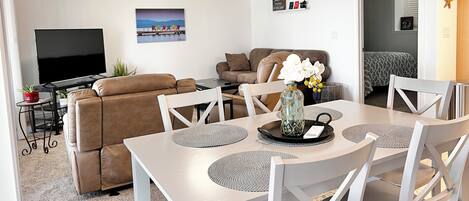 A modern living room with a flat-screen TV, a sofa, a dining table with chairs, and a decorative vase with flowers.