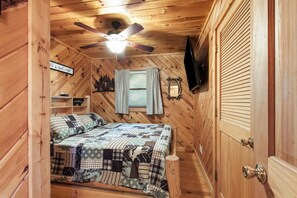 Ein gemütliches Schlafzimmer mit einem Bett, einem Deckenventilator, einem Fenster mit Vorhängen, einem an der Wand befestigten Fernseher und einer Holztür.