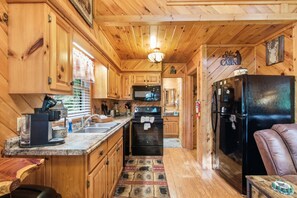 Ein Interieur einer Holzhütte mit Holzwänden, einer Küchenzeile inklusive Herd, Backofen und Kühlschrank sowie einer Sitzecke mit einer Ledersofa.