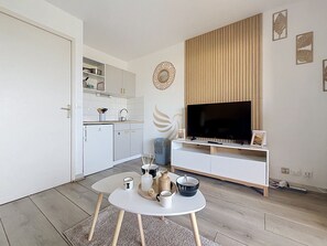 Ein modernes Wohnzimmer mit Fernseher, einem Esstisch mit Tassen und einem Dekorationsobjekt, einer Küche im Hintergrund und einer Wand aus Holzpaneelen.