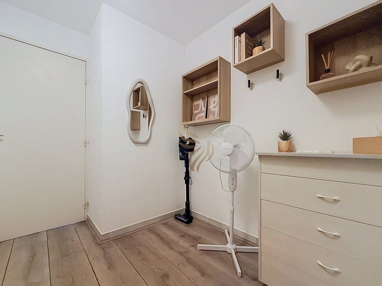 A room with a white door, a standing fan, a wooden shelf with decorative items, and a white dresser with drawers.
