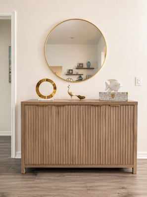 A wooden console with a round mirror and decorative items. - New Designer Home · Complimentary Golf Cart · Prime Location in The Villages (Sumterville)