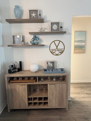 Ein Holzkonsolentisch mit Gläsern und einer Kaffeemaschine, begleitet von Regalen mit Dekoration und einem gerahmten Kunstwerk.