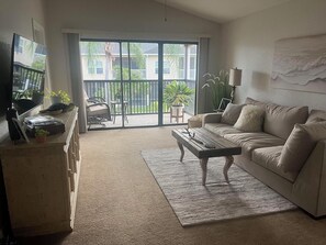 A living room with a sofa, coffee table, and a television on a stand. There is a sliding glass door leading to a balcony with plants and a view of buildings. - NSB Coastal Escape - 3BR Riverside Condo (New Smyrna Beach)