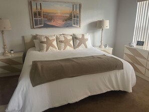 A hotel room with a large bed, starfish pillows, a beach scene painting, a lamp, and a bedside table. - NSB Coastal Escape - 3BR Riverside Condo (New Smyrna Beach)