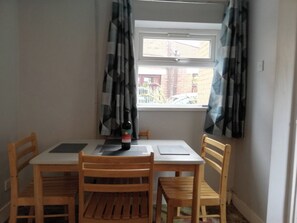 A dining area with a table and four wooden chairs, a bottle of wine, and a window with curtains. - 4 Bedroom House Sleeps 6 - Garden - Free Parking (Belfast)
