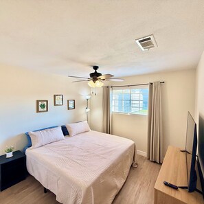 Une chambre à coucher avec un lit, un ventilateur de plafond, une fenêtre avec des rideaux, une table de chevet avec une lampe et un téléviseur posé sur un meuble.