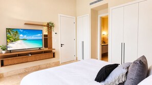 A bedroom with a bed, a TV on a wooden stand, and a view of a beach through the window. - Coastal Voyage 3BR Garden View (Rio Hato)
