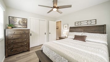 Un dormitorio con un aparador de madera, una cama con ropa de cama estampada, un ventilador de techo y una puerta que da a otra habitación.