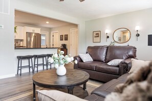 A living room with a brown leather sofa, a coffee table with a vase of flowers, and a kitchen in the background.