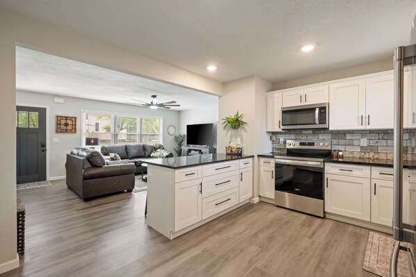 A modern kitchen with white cabinets, stainless steel appliances, and a grey leather sofa in the living area.