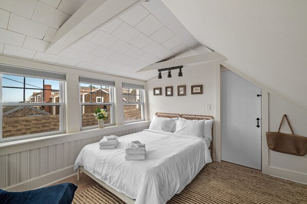 Une chambre avec un grand lit, une fenêtre donnant sur des maisons et une porte à laquelle est accroché un panier.