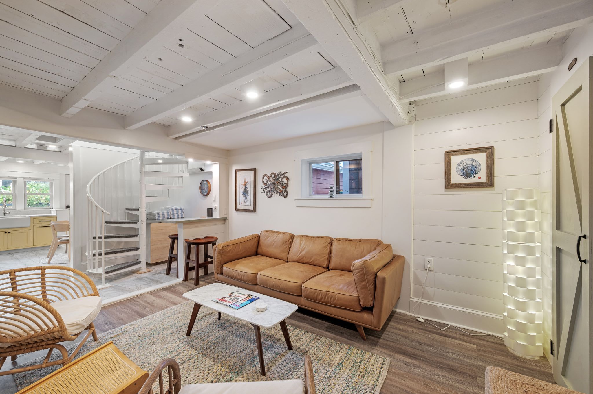 A spacious living area with a brown leather sofa, a white coffee table, and a wicker chair. A staircase leads to an upper level, and a window provides natural light.
