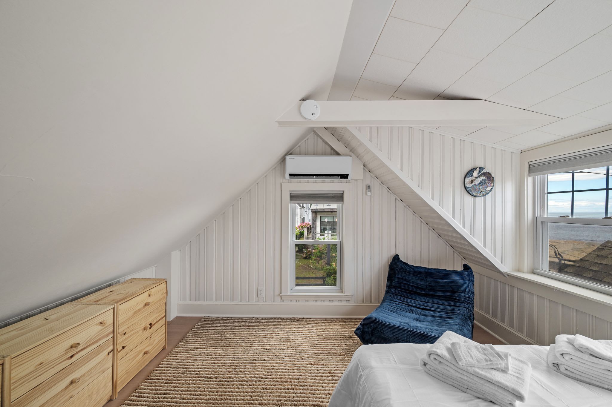 A bedroom with a sloped ceiling, a bed with white linens, a blue pillow, a wooden dresser, and a view through a window.
