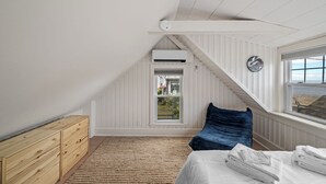 Ein Schlafzimmer mit schrägem Dach, einem Bett mit weißen Leinen, einem blauen Kissen, einer Holzkommode und Blick durch ein Fenster.