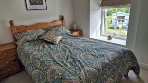 Quarto com cama de madeira, roupa de cama estampada, mesa de cabeceira, abajur, janela com vista para o exterior e quadro na parede.