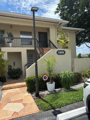 A two-story building with a number '5606' on the facade, a staircase with a railing, and a variety of potted plants and shrubs. - 2-bedroom apartment in peaceful Delray Beach with AC (Delray Beach)